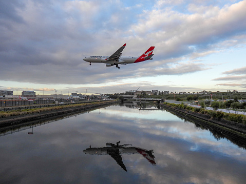 A Qantas Airbus A330-202 plane, registration VH-EBJ, coming into land from the north on the main runway of Sydney Kingsford-Smith Airport as flight QF42 from Jakarta. She is passing over the Alexandra Canal which flows into the Cooks River.  This image faces west and was taken from Nigel Love Bridge, Mascot on a cloudy morning after sunrise on 28 October 2023.