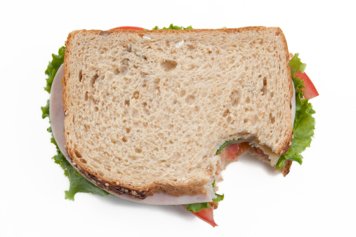 A turkey sandwich on wheat with lettuce and tomatoe sits on a white cutting board.  A bite is taken out of it.  Shallow depth of field.