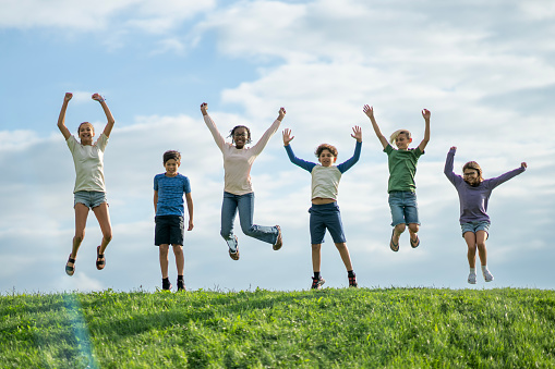 A small group of six elementary aged children stand at the top of a grassy hill, with their hands raised above their heads, as they jump in the air and celebrate the beginning of summer.