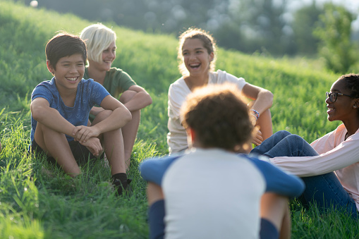 A small group of school aged children are seen sitting in the grass together on a warm summers day.  They are each dressed comfortably as they relax and talk amongst each other.