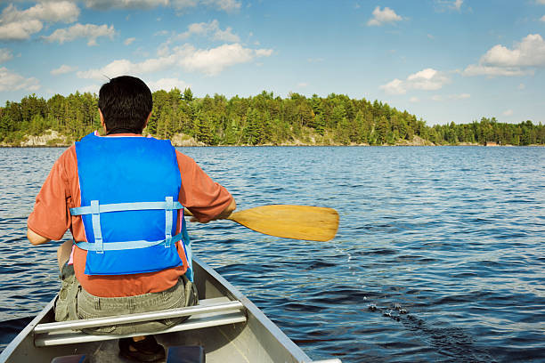 Asian man paddling towards tree shore in aluminum canoe on lake near Boundary Waters Canoe Area, Ely, Minnesota. The vacationing male, wearing life vest and holding wooden paddle, is seated at the vessel front and enjoys his recreational pursuit. Horizontal rear view of unidentifiable person. Blue sky and rippling waves providing copy space. 