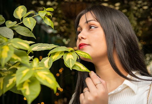 Low angle close up portrait of beautiful serene Indian young woman touching plant’s leafs and contemplating with a smile on her face in nature.