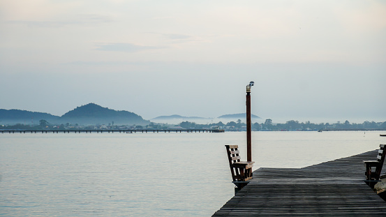 The wooden bridge sits on the edge of a quiet beach, and has a beautiful view of the calm sea water. Wooden bridge as pathway to seaside for docking ship. Minimal sea sky, calm water surface and reflections.