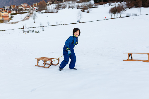 smiling child in blue winter clothes with short dark hair pulling up the wooden sled in snowy mountain