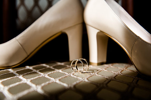 Set against a backdrop of sophisticated geometric patterns, two gold wedding bands take center stage, representing an unbreakable bond of love and commitment. The curves of a pair of white heels frame the scene, casting intriguing shadows that complement the allure of the intertwined rings. The natural light that kisses the surface of the rings creates a dazzling effect, highlighting their circular perfection. This delicate composition captures the essence of matrimonial elegance and the profound symbolism of two lives becoming one.