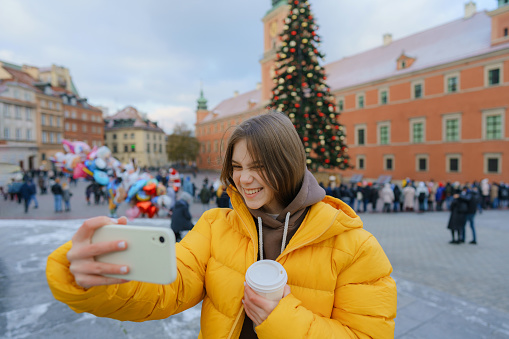 Teenage girl in yellow coat making selfie on  the background of Christmas tree on towns square in Europe
