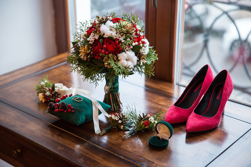 A carefully curated selection of bridal essentials for a winter wedding, showcased atop an antique wooden dresser. A vibrant bouquet, made up of red roses and accented with snow-covered pinecones, sits majestically next to a green velvet box holding wedding rings. A pair of stunning crimson suede heels, placed near a window, completes the scene, hinting at the elegance of the bride.
