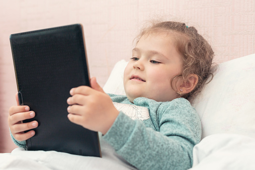 Little girl watching cartoons or playing a game on a tablet in the bedroom at home.
