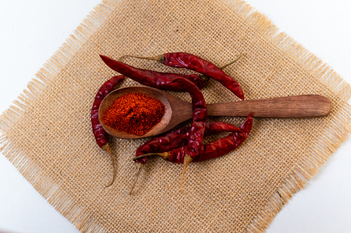 A closeup shot of dried red chili peppers and red chilli powder on wooden spoon, kept on jute fabric background.