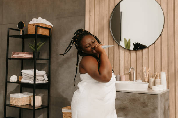 Joyful curvy African woman holding cosmetic bottle like microphone while singing in bathroom