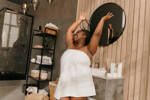 Joyful curvy African woman covered in towel playing with her braided hair and smiling in bathroom