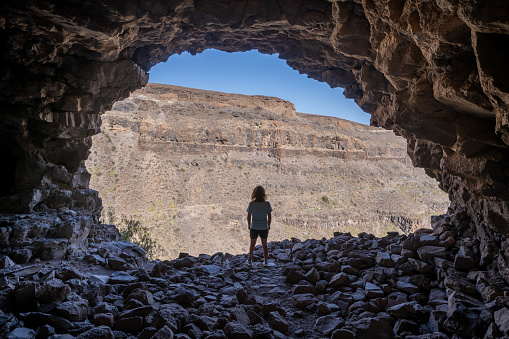 Rear view of a woman standing in the access of a large cave looking outside in the archaeological complex 