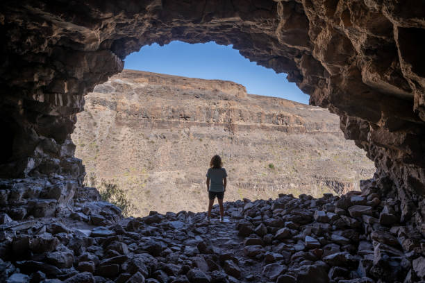 Rear view of a woman standing in the access of a large cave looking outside in the archaeological complex "La Fortaleza", in Santa Lucía De Tirajana, on the island of Gran Canaria, Spain stock photo
