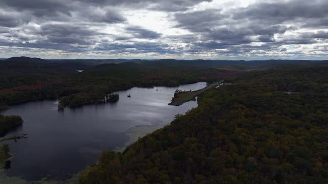 Aerial view of the countryside in Stormville, New York on a cloudy day in the fall with Black Pond in view. The camera dolly in then truck right along the pond towards an abandoned aircraft runway.