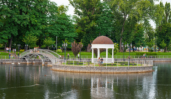 Kremenchuk city, Ukraine - July 26, 2022: A beautiful pond with a white gazebo on an island and an iron pedestrian bridge. City garden with tall trees on a summer day