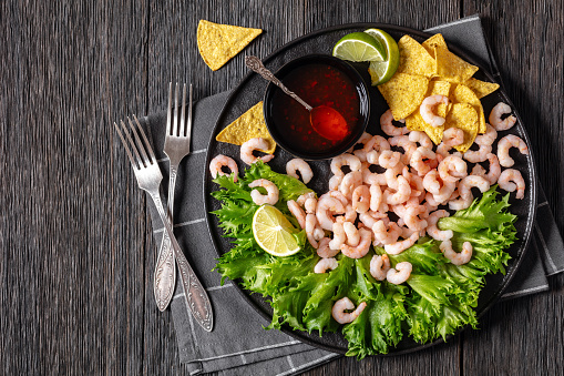 steamed shrimps with lettuce, lime wedges, corn chips and sweet chili sauce on black plate on dark wooden table, horizontal view from above, flat lay, free space
