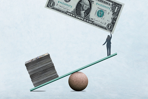 A woman reaches for a one dollar bill as she stands at the end of a seesaw that is lifted by a tall stack of money on the other end illustrating the concept that it takes money to make money.