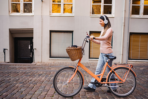 Brunette woman with white wireless headphones listening to music while walking with bicycle through the city