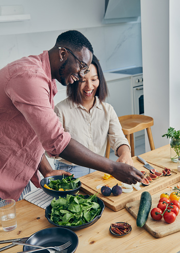 A beautiful & happy mixed-race couple, asian and black, in the modern white kitchen, enjoying a brunch preparation through positivity and happiness, sharing and representing love, joy, wellbeing, true couple friendship, security, trust and togetherness