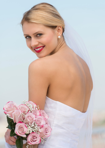 Rear view portrait of beautiful young bride in backless wedding dress holding flower bouquet against sky