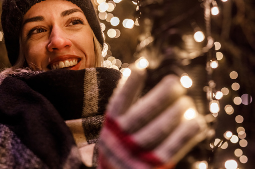 Close up shot of cheerful young woman standing by the pole wrapped in glistening Christmas lights, looking away, smiling and contemplating.
