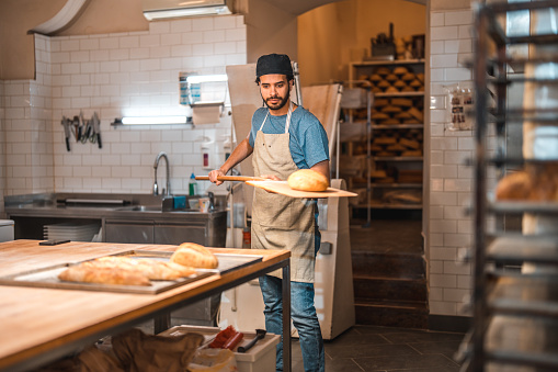 Middle Eastern male baker taking out a freshly baked bread out of the oven. He is using a shovel and enjoys working in an artisan bakery.