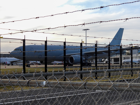 A Royal Australian Air Force Airbus KC-30A, the military version of an Airbus A330-200, registration A39-007, parked at Sydney Kingsford-Smith Airport.  She returned from Washington earlier in the day, having transported the Prime Minister to meet with President Biden.  In the background is a Virgin Australia B737 plane.  This image faces west and was taken from Ross Smith Avenue, Mascot on a sunny afternoon near sunset on 28 October 2023.