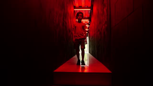 Young boy moves through dark corridor illuminated by haunting red light