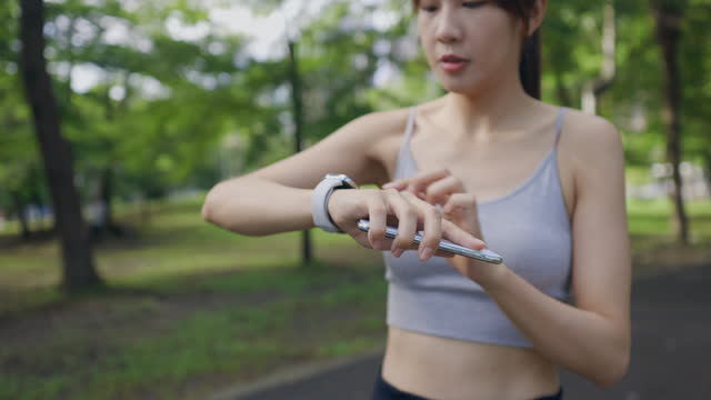 Ａn Asian woman is seen attentively reviewing the fitness app on her phone after finishing her jogging training. She meticulously compares the data on her fitness watch to confirm the effectiveness of her workout and ensure she has achieved her goals.