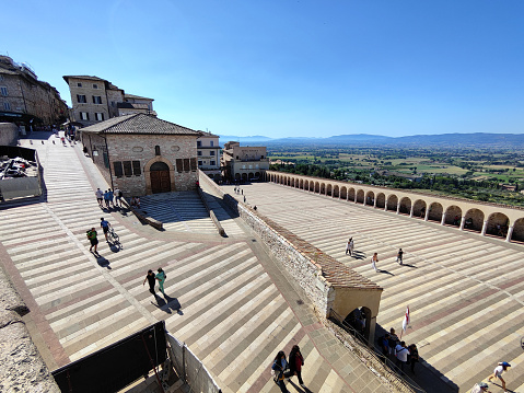 Tourists walking at the lower square at the Basilica di San Francesco d'Assisi, a town in the Umbria region in central Italy, where Saint Francis was born and died. It is one of the most important places of Christian pilgrimage in Italy. The basilica is a distinctive landmark and has been a UNESCO World Heritage Site since 2000.