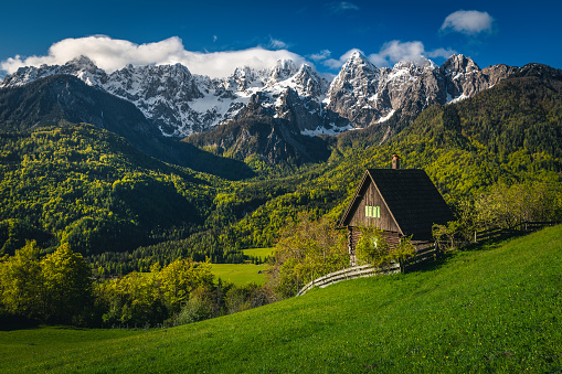 Amazing spring alpine scenery with cute wooden hut on the green field and high snowy mountains in background, Srednji Vrh village, Kranjska Gora, Julian Alps, Slovenia, Europe