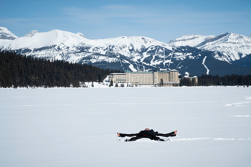 Woman makes a snow angel in the snow on frozen Lake Louise in front of Fairmont Chateau and the mountains of Lake Louise Ski Resort in the distance, Banff National Park, Alberta, Canada