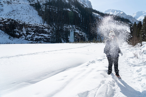 A female wearing crampons over her winter boots throws snow at someone while hiking at Lake Louise in winter, Banff National Park, Alberta, Canada
