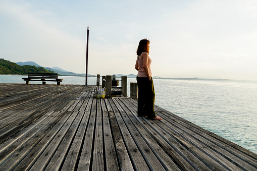 Asian women stand on a wooden pier with her back. Woman enjoying the sunrise. Silhouette alone woman relax watching sunrise and cloud sky. Thinking and concentrate in summer background with copy space for label text.