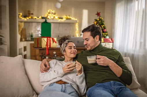 Young couple sitting on sofa at home and enjoying in their time together. They are drinking coffee. It is Christmas time.
