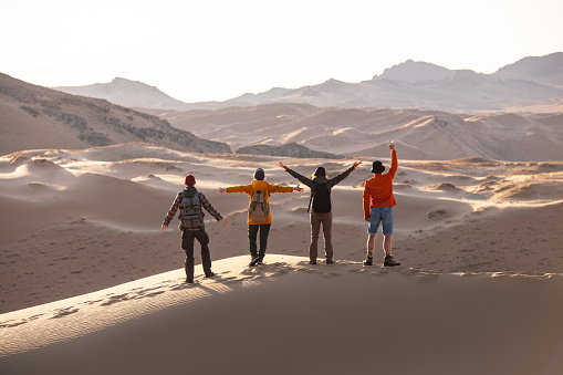 Four happy tourists with backpacks stands on sandy dune with open arms and looks at sunset