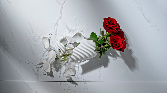 Overhead view of broken vase with red rose flowers on white marble floor.