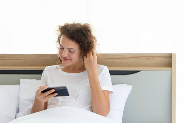 Junge schöne glückliche Frau, die sich entspannt und Smartphone benutzt, während sie morgens nach dem Aufwachen im Bett liegt. – Foto