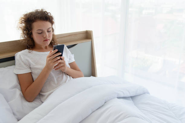 Joven hermosa mujer feliz relajándose y usando el teléfono inteligente mientras está acostada en la cama por la mañana después de despertarse. - foto de stock