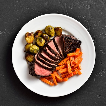 Grilled beef steak with brussels sprouts and sweet potatoes on plate over black stone background. Top view, flat lay