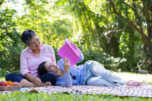 アジアの娘 母親の膝の上で眠り、公園のピクニックマットの上で本を読んでいます。幸せそうな先輩女性が娘と話す。医療・介護支援サービスのコンセプト - grandparent senior adult child reading ストックフォトと画像
