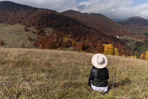 Young woman sitting in a meadow and admiring the beautiful autumn landscape in front of her eyes.