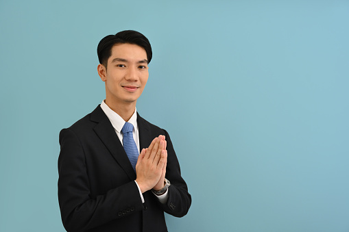 Waist up Portrait studio shot of a Young friendly Asian man in an office business suit standing with a smile face posing hands for greeting or thank you in Asian style over a color isolated background.