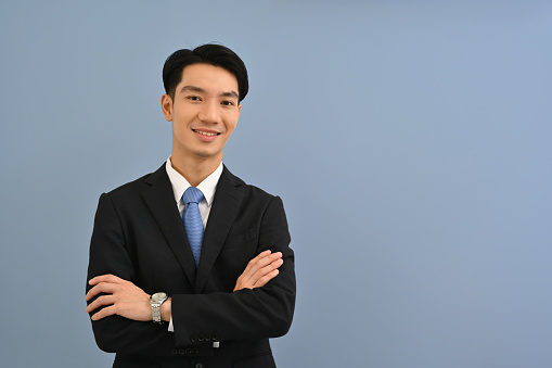 Waist up portrait of a Confident Young Asian Businessman in a formal suit standing with arms crossed gesture over a color isolated studio background.