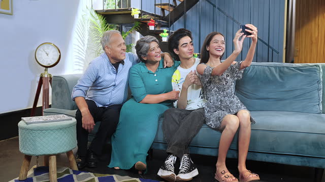 Happy Indian excited family sitting on sofa smiling teenage girl hold smartphone taking selfie at house. Old aged grandparents couple make funny pose photo or record video blog enjoy weekend together