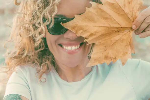 Playful happy woman portrait smiling and hidden half face with big autumn dry yellow maple leaf. Cheerful young adult female people smile and look on camera wearing sunglasses in outdoor leisure day