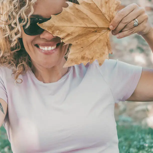 Playful happy woman portrait smiling and hidden half face with big autumn dry yellow maple leaf. Cheerful young adult female people smile and lok on camera wearing sunglasses in outdoor leisure day