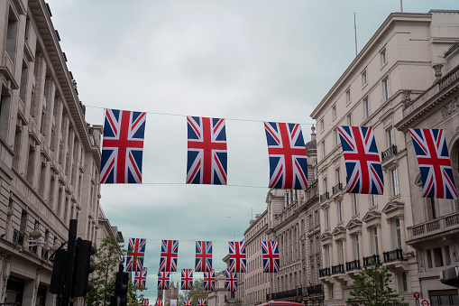 London, UK - May, 2023. People crossing street and sit on steps of monument at Piccadilly Circus Street in London during daytime in springtime with Piccadilly Circus Shopping center behind and huge advertisement board. British Flag and Coronation Flag  Landscape format.