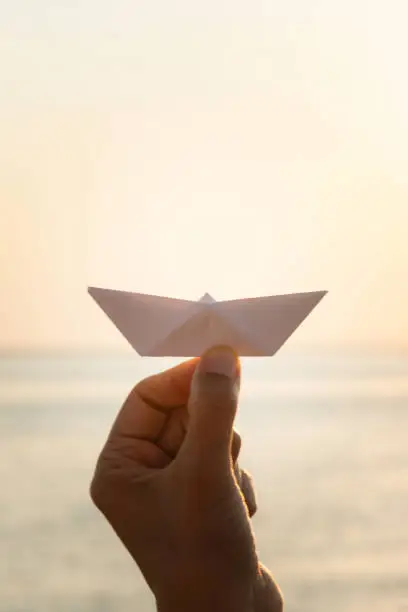 Hand of unrecognizable caucasian female at the sea  is showing a white paper boat at camera.