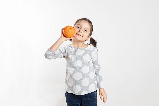 Girl is holding an orange in front of a white wall.